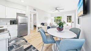 Una cocina moderna con armarios blancos, un refrigerador de acero inoxidable y una cafetera. Hay un comedor con una mesa redonda y sillas azules.