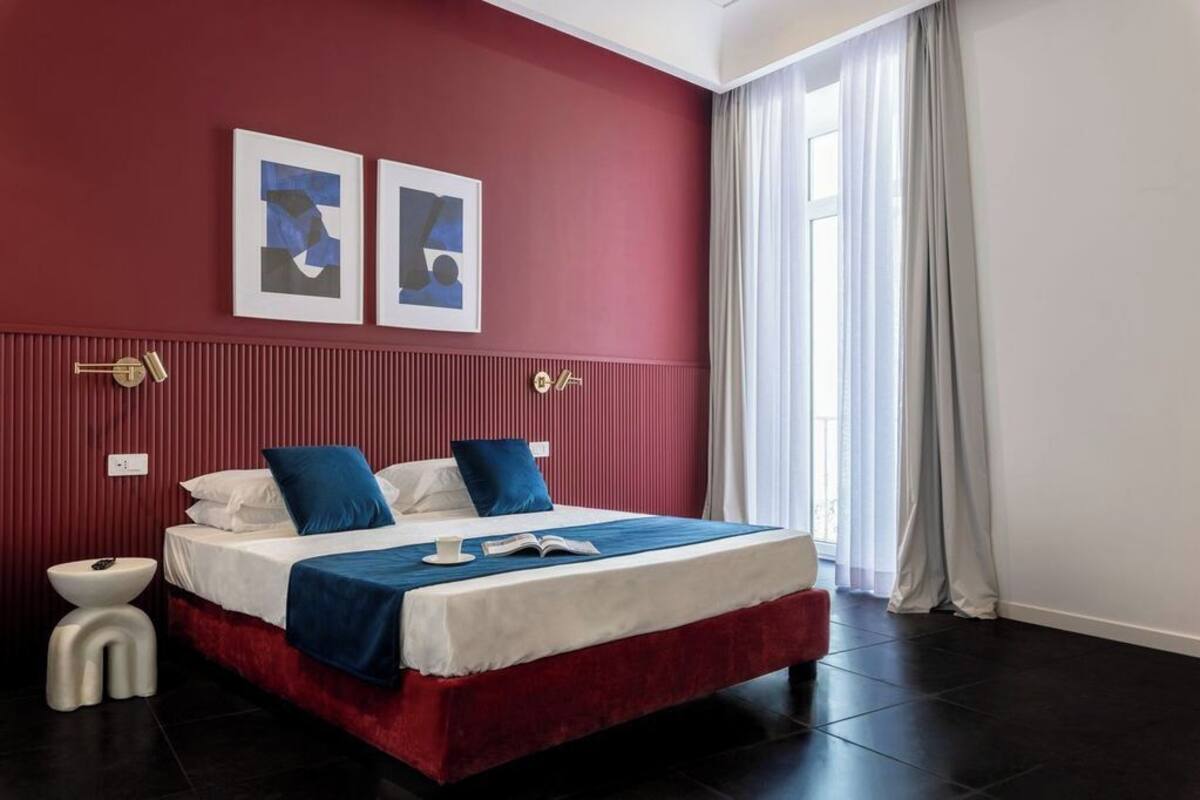 Un dormitorio moderno con una pared roja de acento, una cama grande con ropa de cama azul, una mesita de noche y una ventana con cortinas.