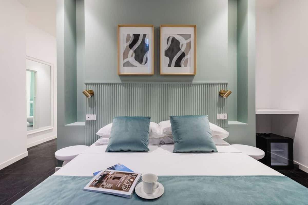 Una habitación de hotel moderna con una cama, dos almohadas, una mesita de noche con una revista y una taza, y dos cuadros abstractos en la pared.