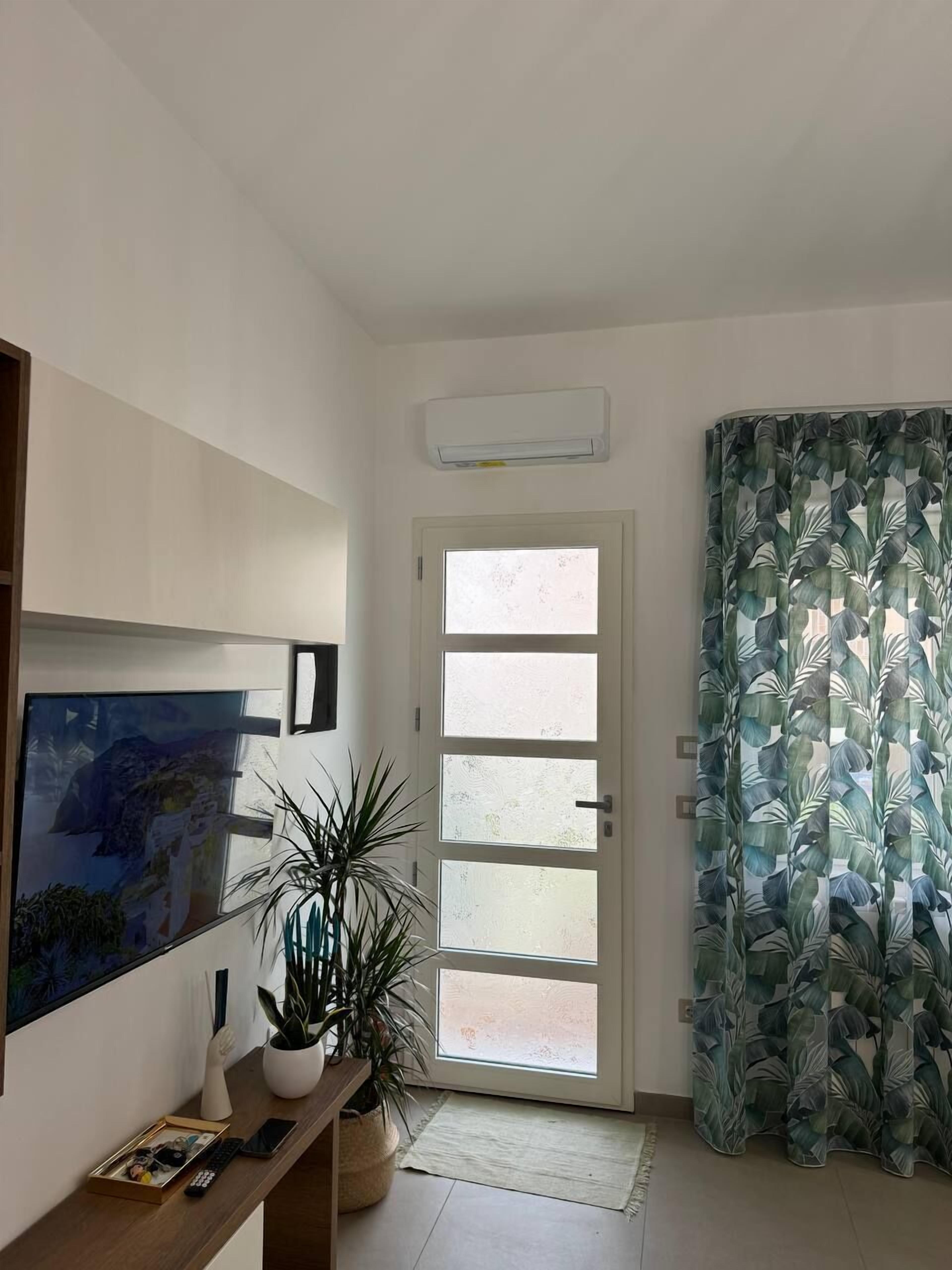 A room with a large window, a potted plant, a wall-mounted air conditioner, and a framed picture.