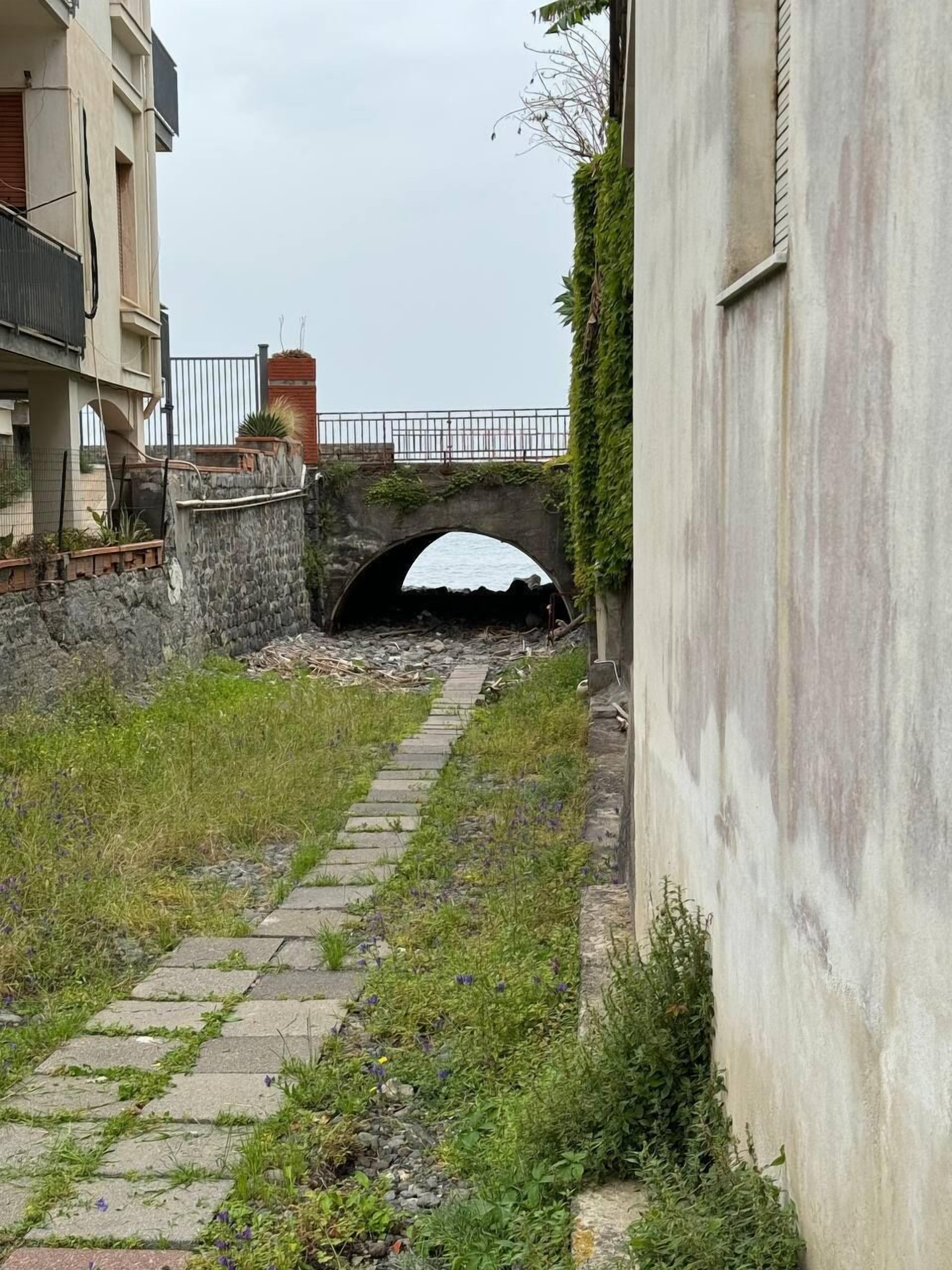Un estrecho sendero de piedra que conduce a un puente de piedra sobre un cuerpo de agua, rodeado de vegetación y un edificio con cerca de metal.