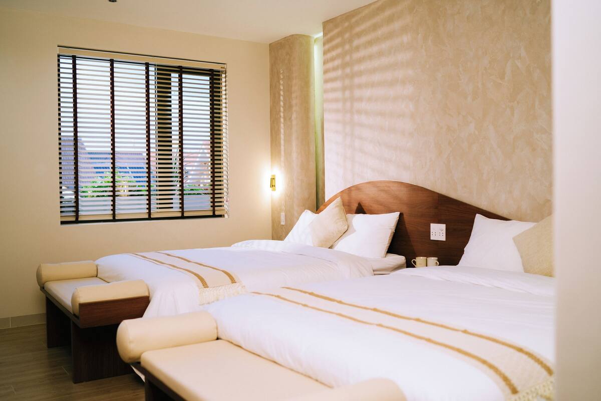 Habitación de hotel con dos camas, un ventanal grande con persianas, un cabecero de madera y una mesita de noche con una taza.