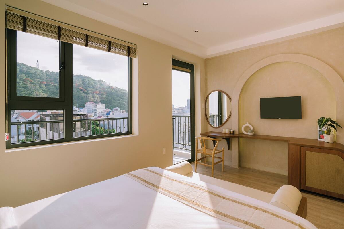 Un dormitorio con una cama grande, un escritorio, una silla, un televisor y vistas de un paisaje urbano a través de amplias ventanas.