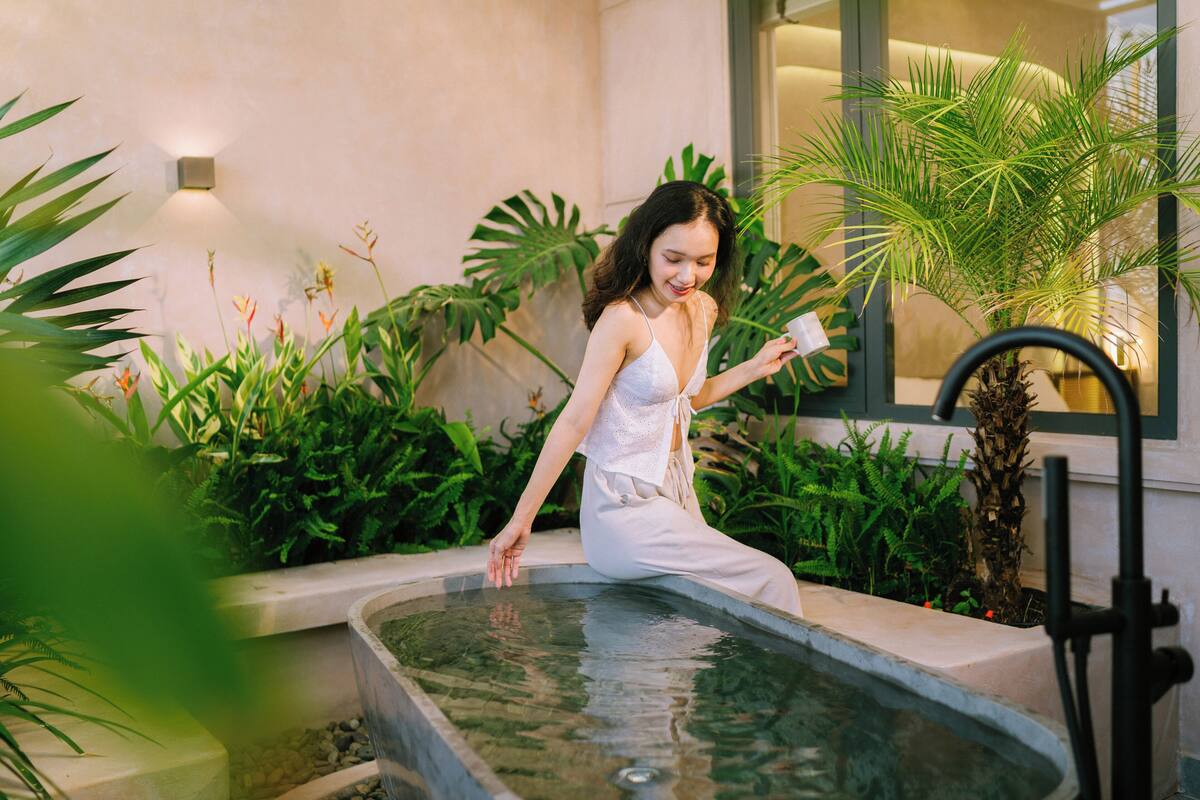 Una mujer con un vestido blanco sentada junto a una piscina pequeña rodeada de abundante vegetación.