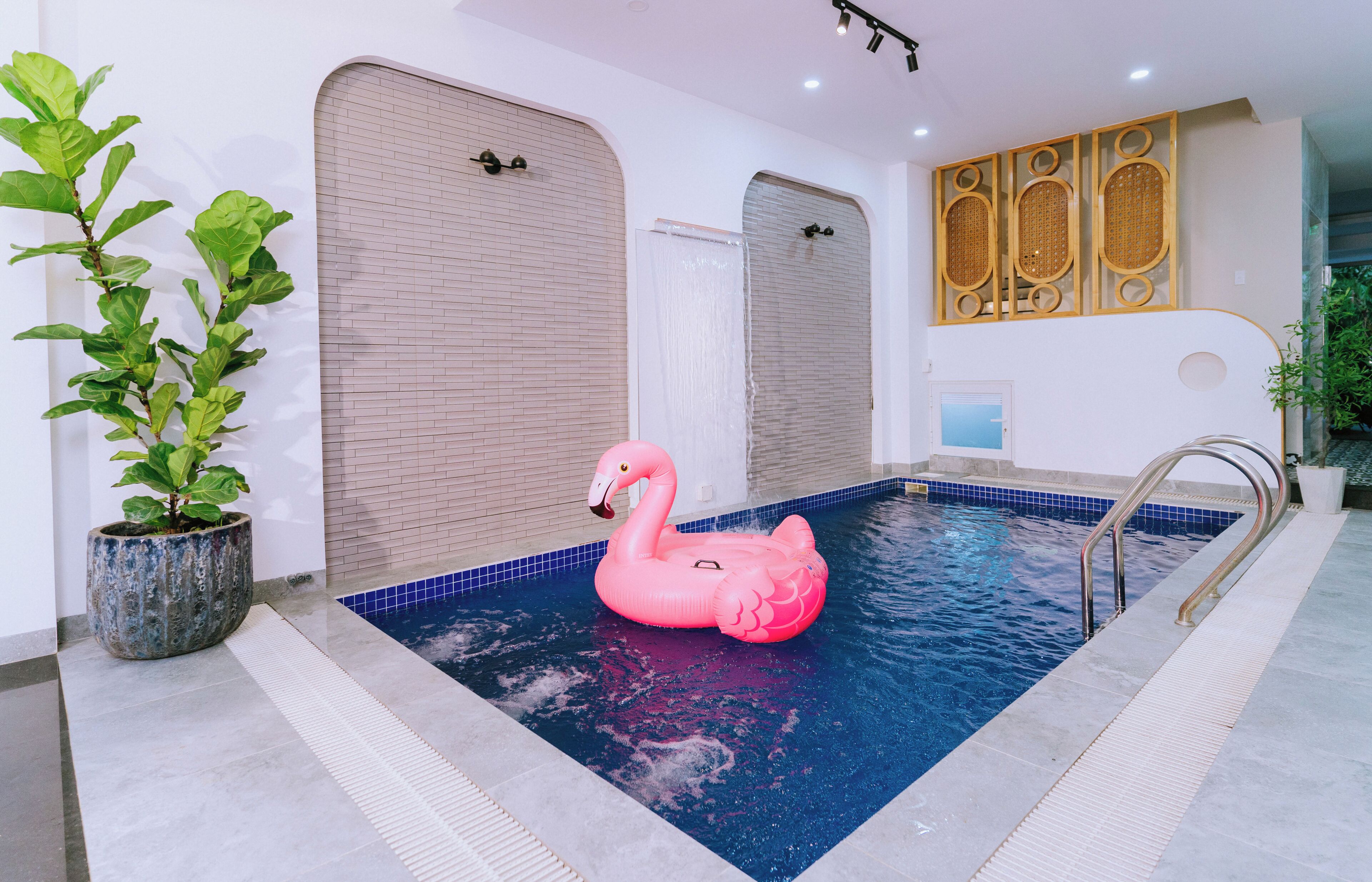 An indoor swimming pool with a pink flamingo float, a potted plant, and a tiled wall.