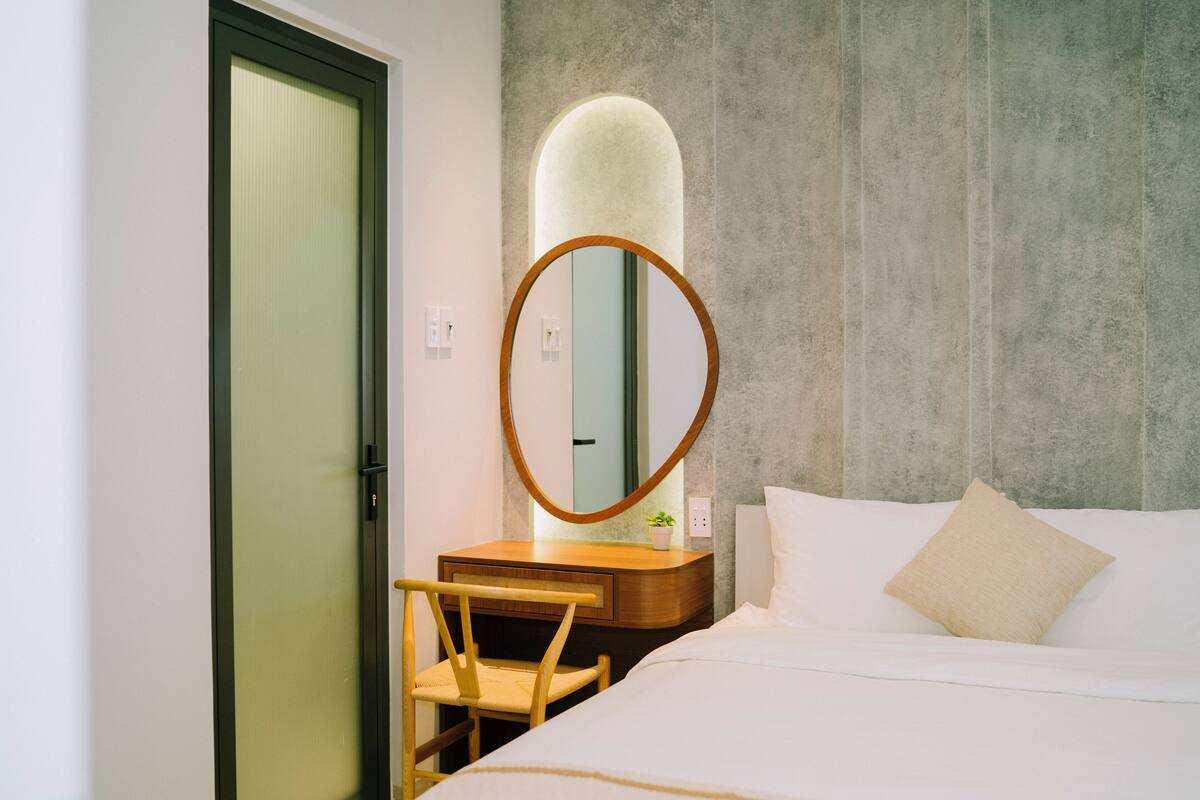 Habitación de hotel con cama, mesita de noche de madera, espejo redondo, planta pequeña en maceta y puerta con marco verde.