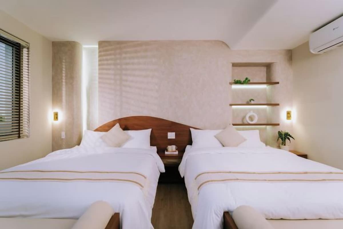 A hotel room with two beds, a wooden headboard, bedside tables with lamps, and a wall-mounted shelf with decorative items.