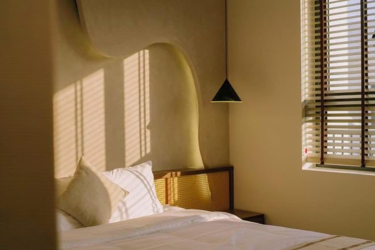 A bedroom with a bed, a bedside table, a lamp, and a window with blinds.