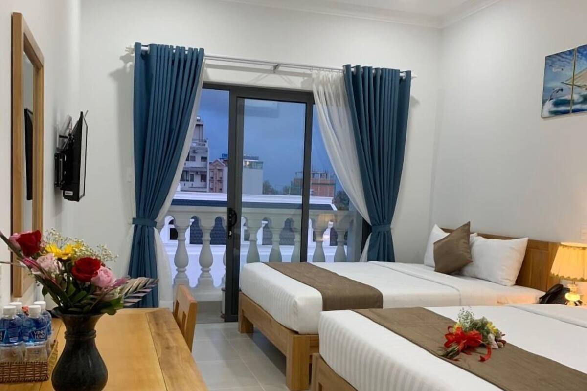 Ein Hotelzimmer mit zwei Betten, einem Holztisch mit Blumenvase und einem Balkon mit blauen Vorhängen.