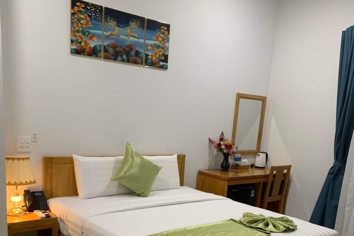 Ein ordentlich bezogenes Bett mit einer grünen Tagesdecke, ein Nachttisch mit einer Lampe, ein Spiegel und ein kleiner Tisch mit einem Blumenstrauß.