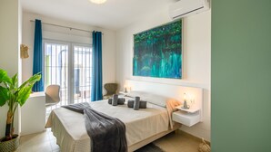 A modern bedroom with a large bed, a desk, a chair, a potted plant, and a large painting on the wall.