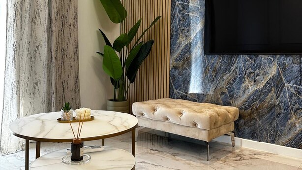 A modern living room with a marble floor, a round coffee table with a candle, a tufted bench, a large plant, and a TV mounted on a marble wall. - Apartment Ivan With Pool (Zagvozd)