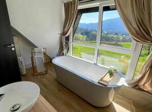 Ein modernes Badezimmer mit freistehender Badewanne, einem großen Fenster mit Blick auf eine grüne Landschaft und einem Holzboden.