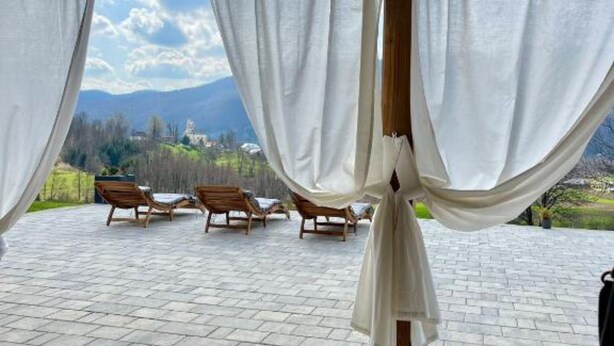 A covered outdoor seating area with wooden loungers and white curtains, overlooking a mountainous landscape.