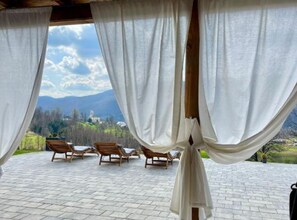 A covered outdoor seating area with wooden loungers and white curtains, overlooking a mountainous landscape.