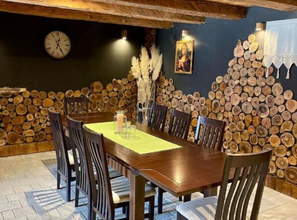 A dining area with a wooden table and chairs, a clock on the wall, and a stack of logs against the wall.