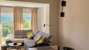 A modern living room with a sofa, a round table, and a view of a forest through large windows.