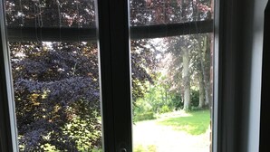 Näkymä ikkunasta, jossa on metallinen kaide, ja ikkunasta avautuu puutarha, jossa on nurmikkoa ja puita.