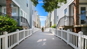 Une terrasse en bois avec des balustrades blanches, bordée de maisons en briques et en bardage, qui mène à l’entrée principale.