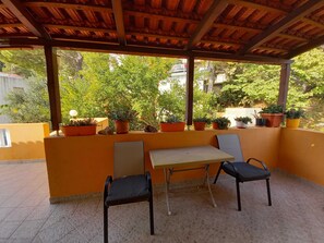 Une terrasse couverte avec une table et deux chaises, entourée de plantes en pot et offrant une vue sur la verdure.