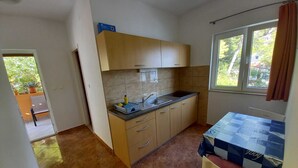 Eine kleine Küche mit Spüle, Arbeitsplatte und Schränken. Ein Fenster mit Vorhängen bietet Blick ins Grüne.