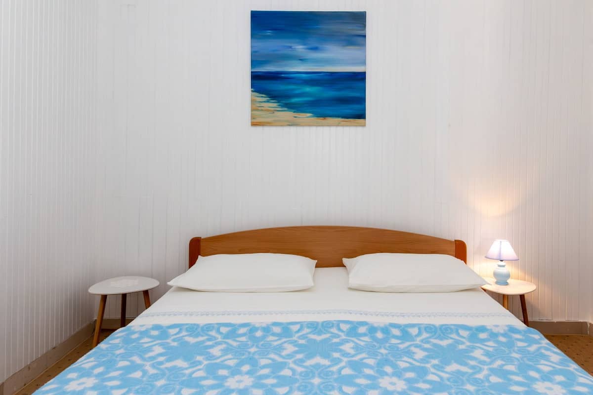A bedroom with a wooden headboard, two white pillows, a patterned blue bedspread, a small bedside table with a lamp, and a painting of a blue seascape on the wall.