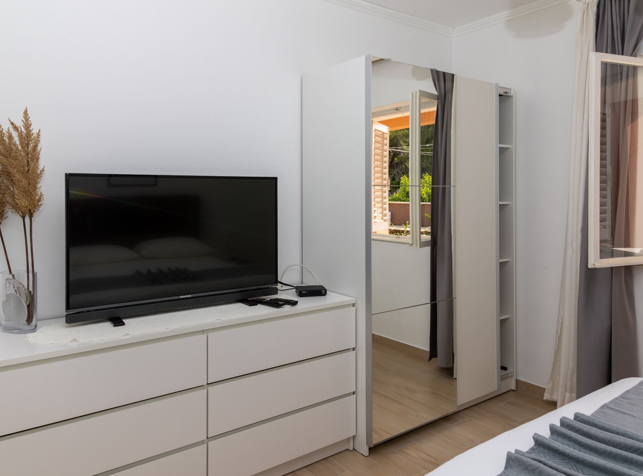 A modern bedroom with a flat-screen TV on a white dresser, a mirror, and a view of a balcony.