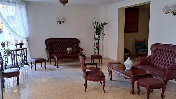 Un salón amplio con un sofá grande, dos sillones, una mesa de centro y una mesa de comedor. Hay una planta en maceta y una máquina de coser sobre una mesita auxiliar. El salón tiene un techo alto con una araña y un ventanal con cortinas.
