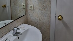 Baño con lavamanos blanco, espejo, inodoro y zona de ducha. Hay repisas con artículos de aseo y una lámpara sobre el espejo.