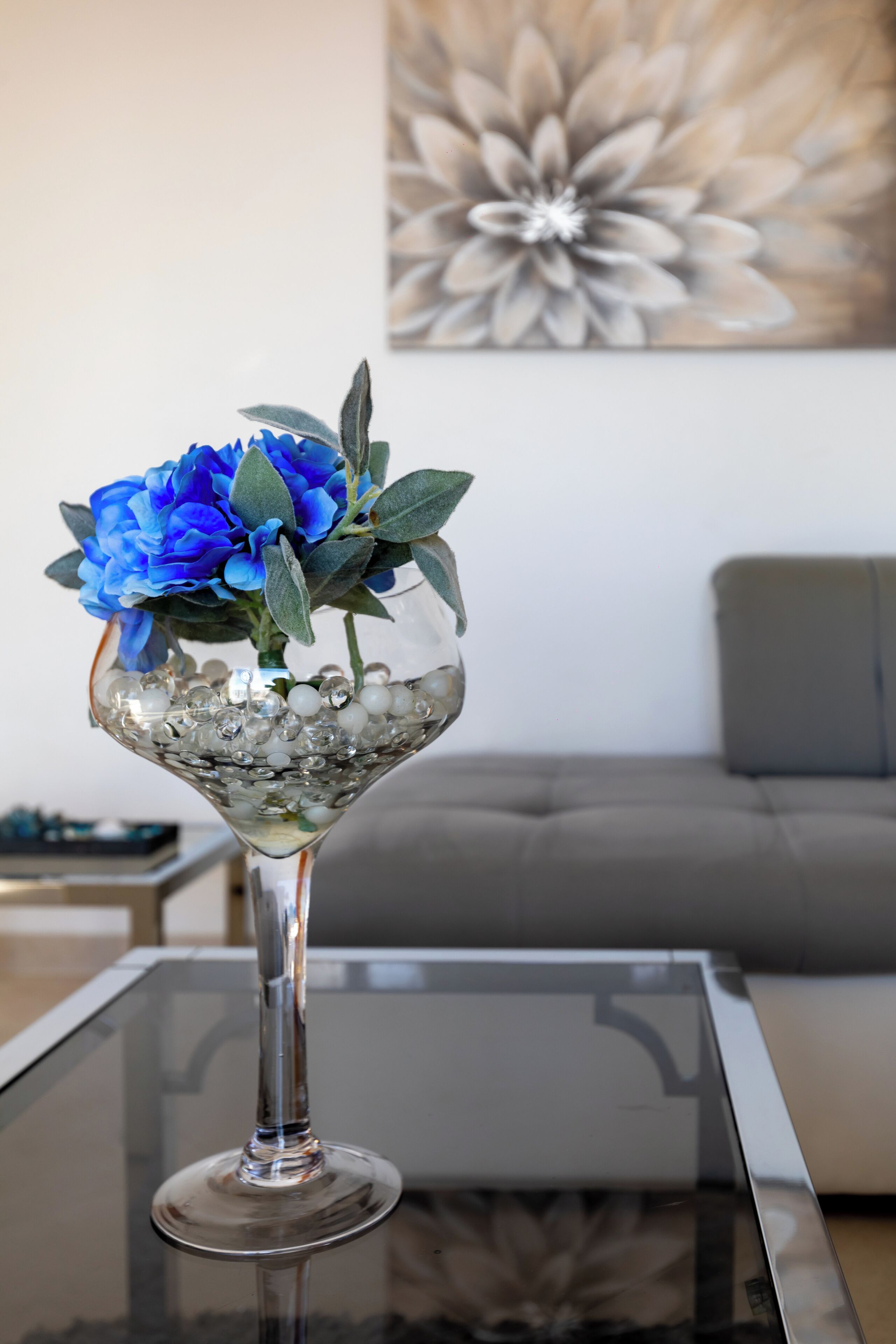 Un vase en verre transparent contenant des fleurs bleues et des feuilles vertes, posé sur une table en verre.
