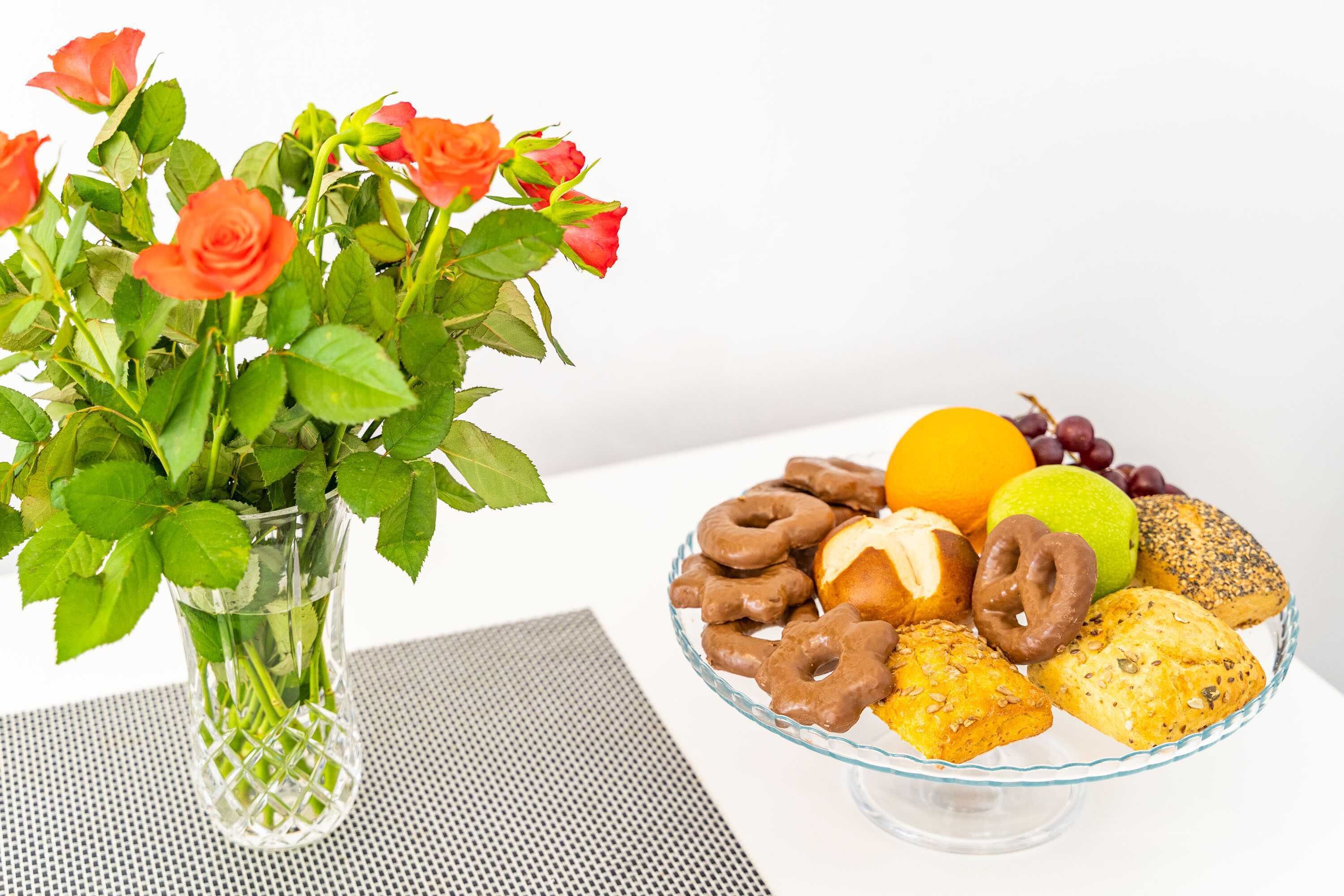 Un vase en verre contenant des roses et un saladier en verre rempli d’une variété de pâtisseries et de fruits.