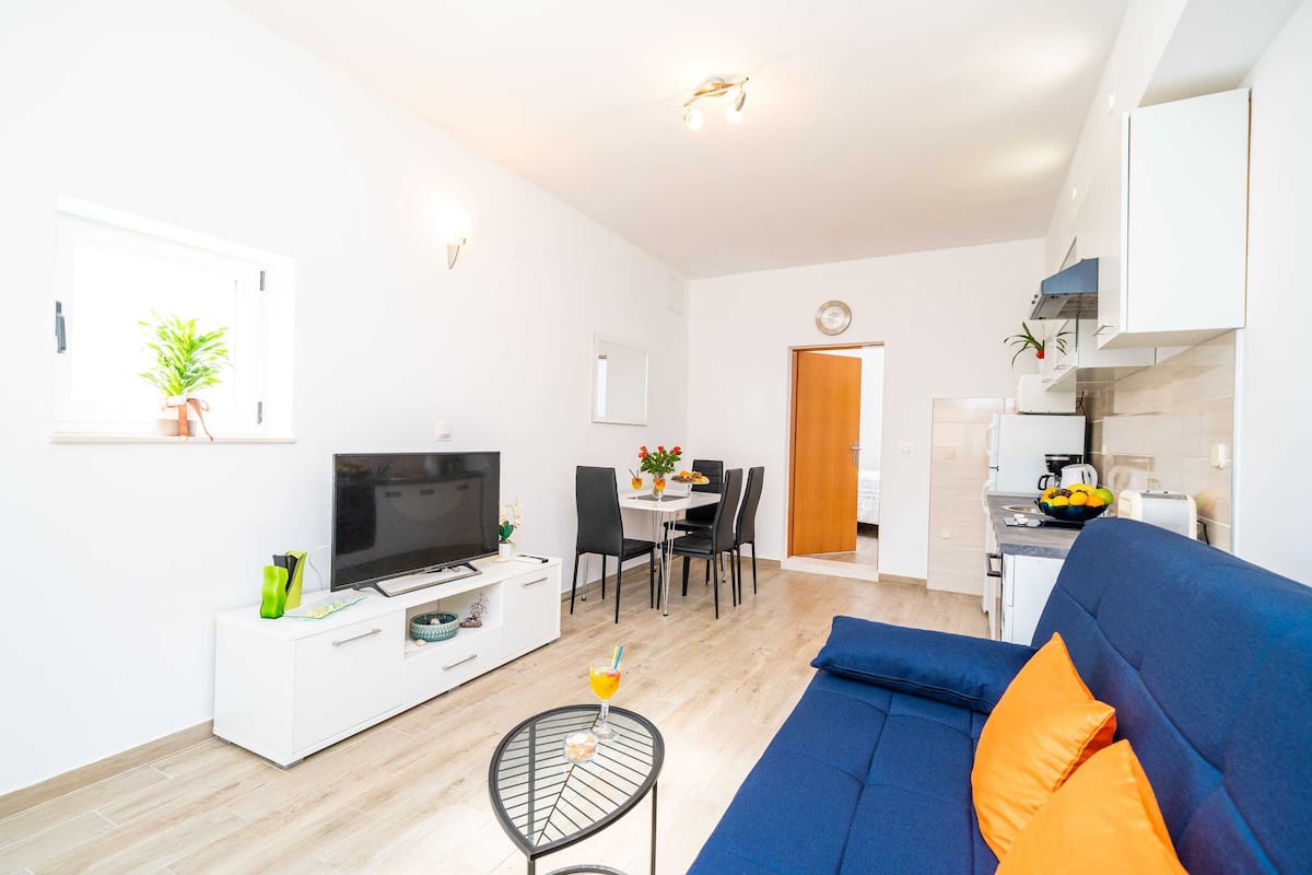 Uma sala de estar moderna com um sofá azul, uma televisão, um espaço de jantar com mesa e cadeiras, e uma cozinha com fogão e pia.