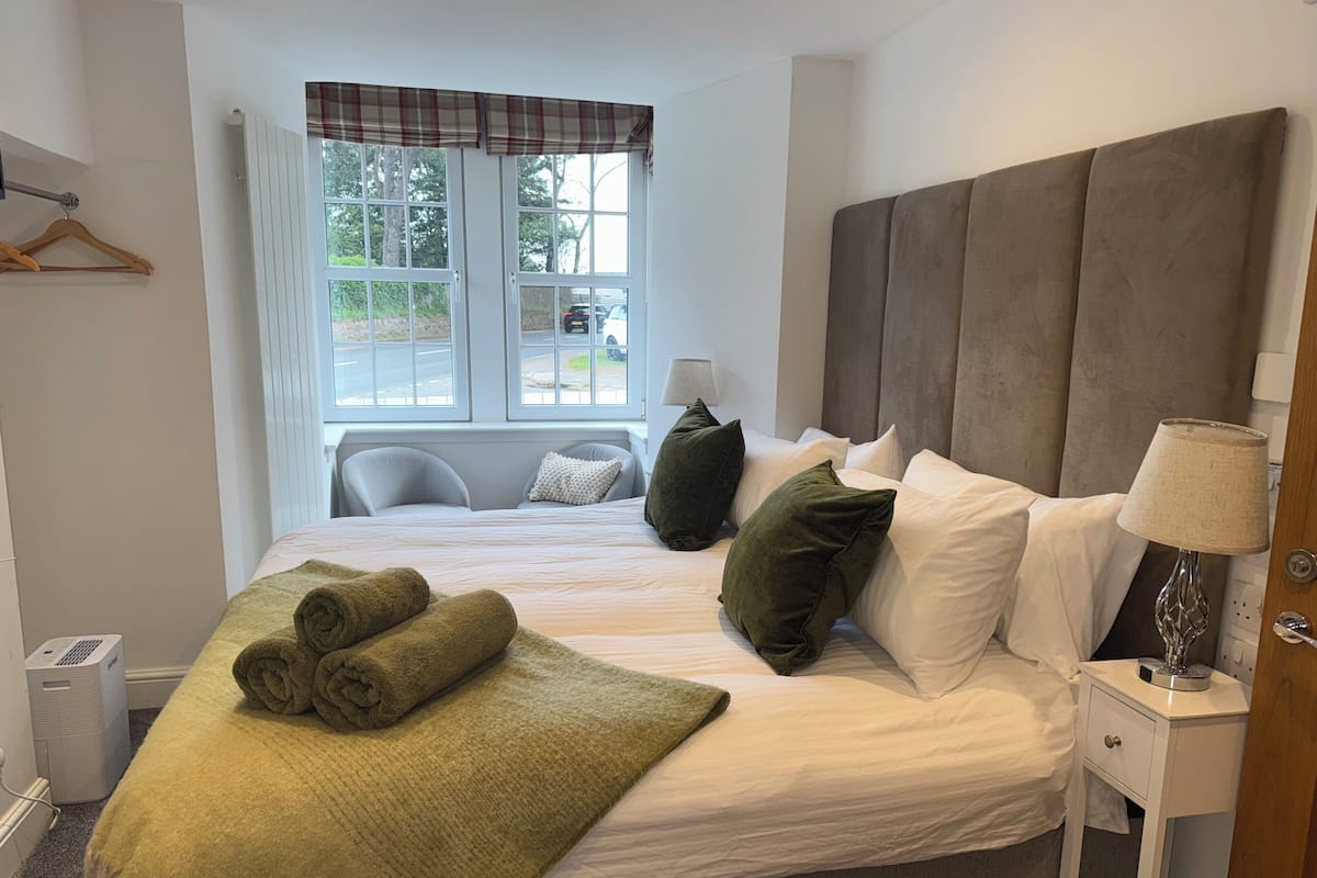 A neatly made bed with pillows and a blanket, a bedside lamp, and a window with a view of the outside.