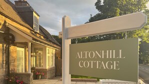 Señal de Altonhill Cottage, con fachada de piedra, techo de paja y jardín bien cuidado.