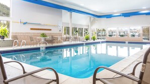 En indendørs swimmingpool med klart, blåt vand, omgivet af liggestole og borde, med store vinduer der giver udsigt til bygninger og træer.