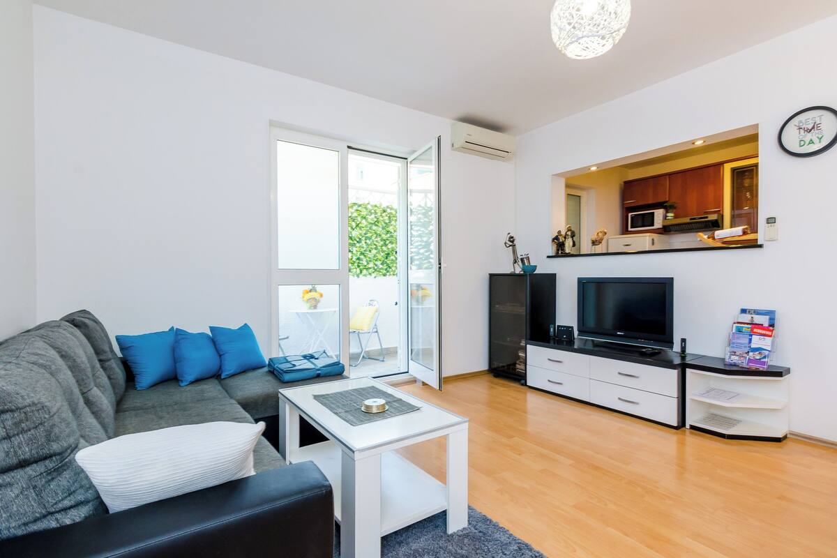 A modern living room with a grey sofa, a white coffee table, a flat-screen TV, and a kitchen area visible through an open door.