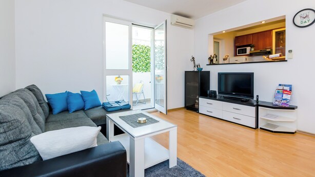 Ett modernt vardagsrum med en grå soffa, ett vitt soffbord, en platt-TV och ett köksutrymme som syns genom en öppen dörr.