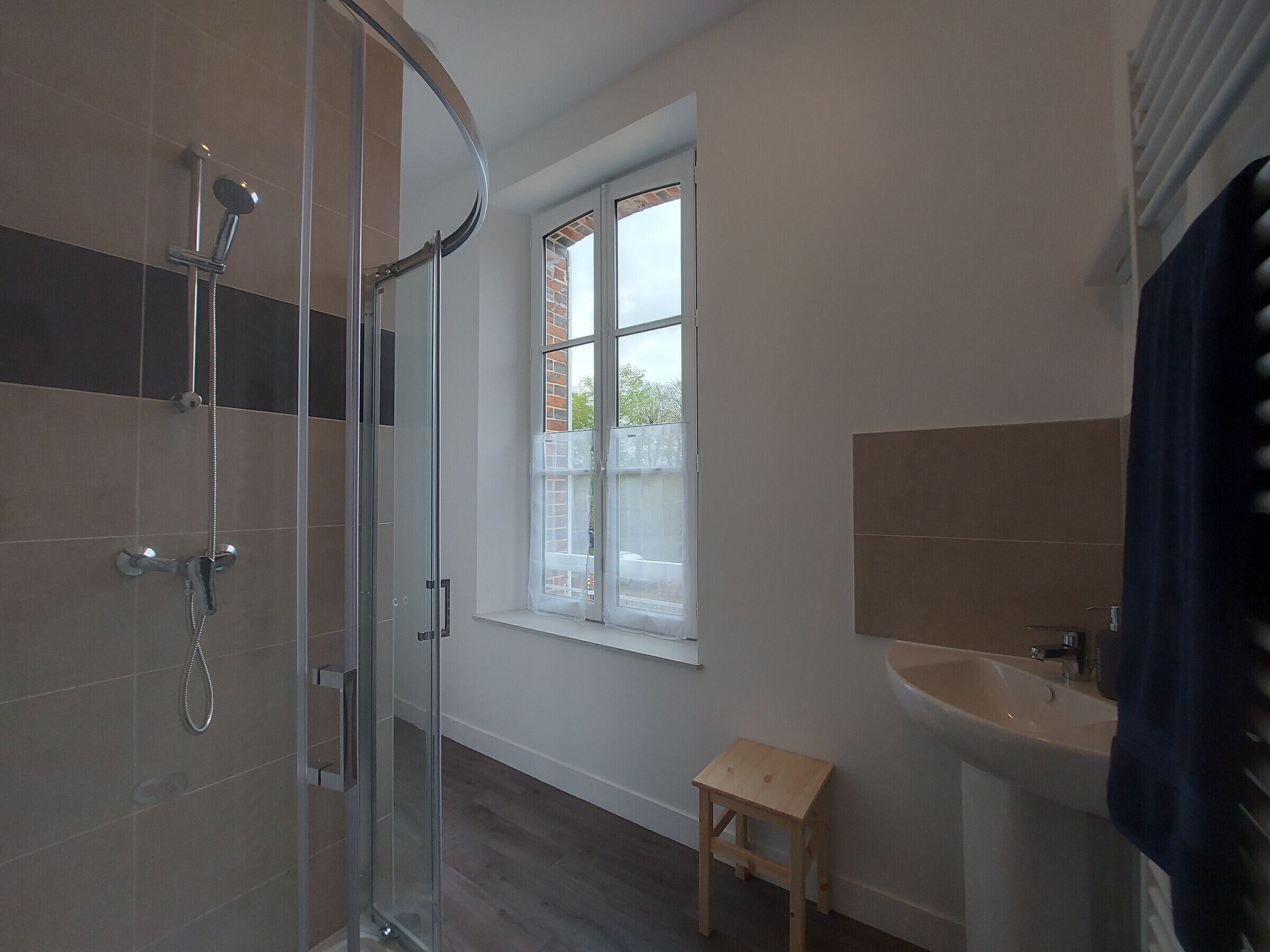 A modern bathroom with a glass shower enclosure, a small wooden stool, and a window providing natural light.
