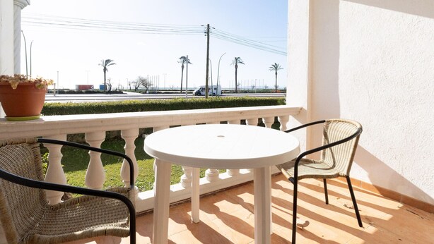 A balcony with a white table and chairs, a potted plant, and a view of the street and palm trees.