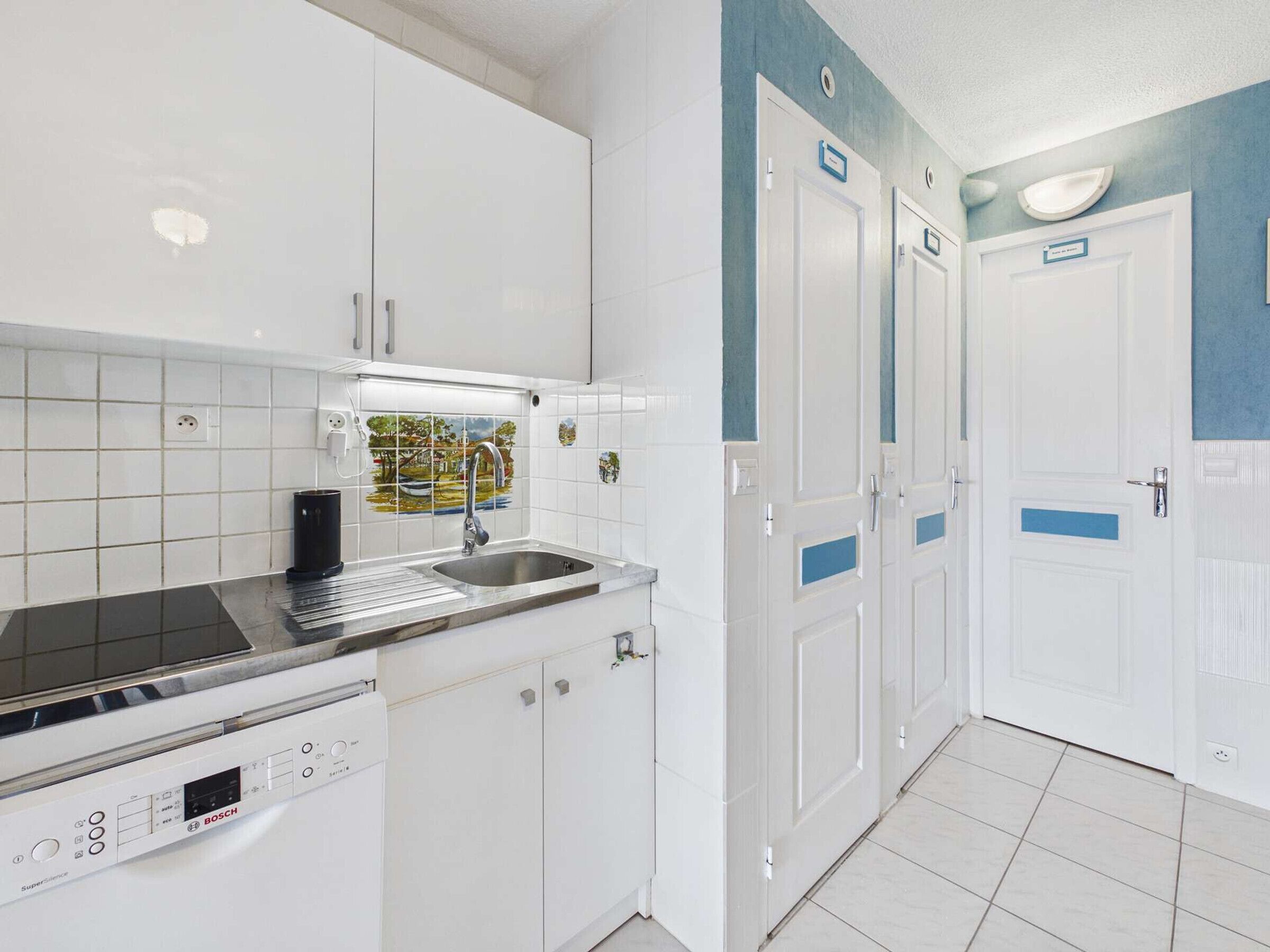 Una cocina con armarios blancos, un lavavajillas Bosch, un lavabo con grifo y salpicadero de azulejos. Hay tres puertas con detalles azules y un interruptor de luz en la pared.
