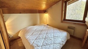 A bedroom with a wooden ceiling, a large window, a bed with a quilted duvet, a bedside table, and a wall-mounted light. - Charming 2-BR + Cabin, Sleeps 6, Close to Jaillet Gondola and Sports Palace - Megève (Megève)