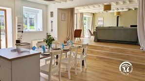 A spacious living area with a wooden floor, a dining table set for four, and a sofa in the background. - Comfortable country cottage - 6 guests (Mayenne)