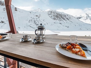 A wooden table with a plate of pizza, a coffee pot, and cups, set against a snowy mountain backdrop.
