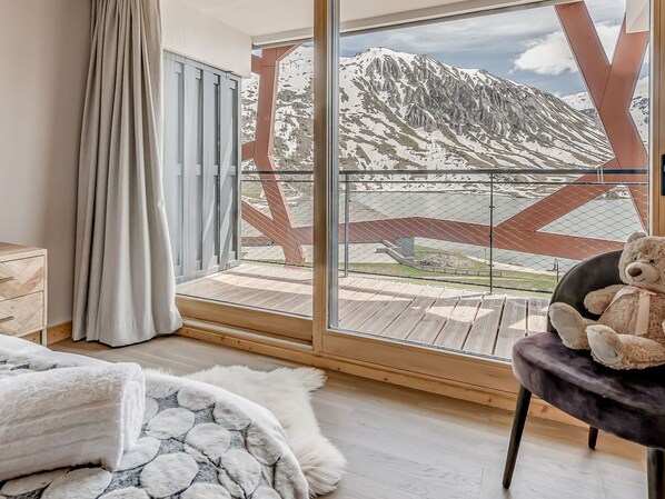 A room with a large window, a wooden floor, a bed with a white blanket, a chair with a teddy bear, and a view of a mountain. - Premium 4* Apartment 8 Pers, Lake View, Wifi, Tignes (Tignes)