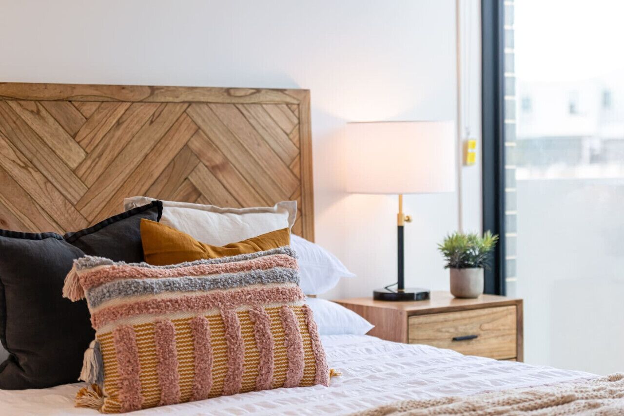A neatly made bed with a variety of pillows, a wooden headboard, a bedside lamp, and a small plant on a nightstand.