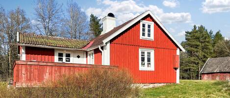 A red wooden house with a sloped roof and a chimney, surrounded by greenery and trees.