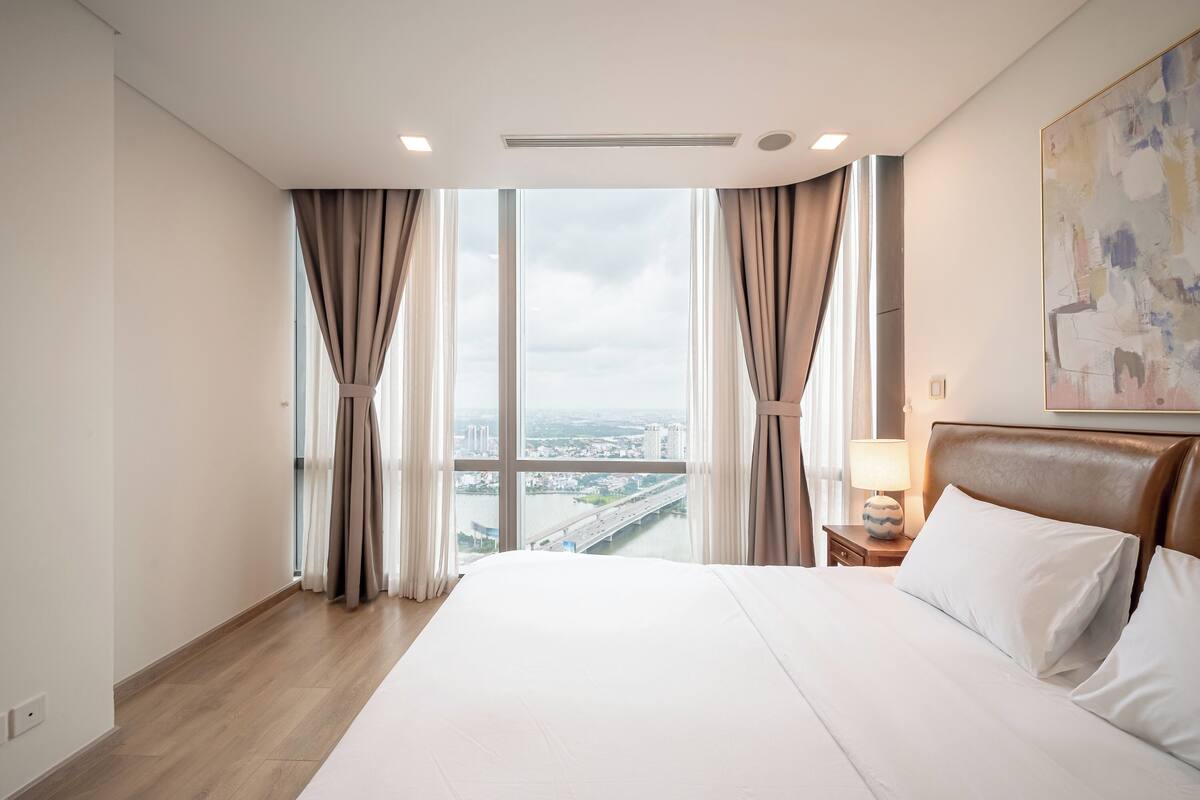 A hotel room with a large bed, a nightstand with a lamp, and a view of a cityscape through large windows.