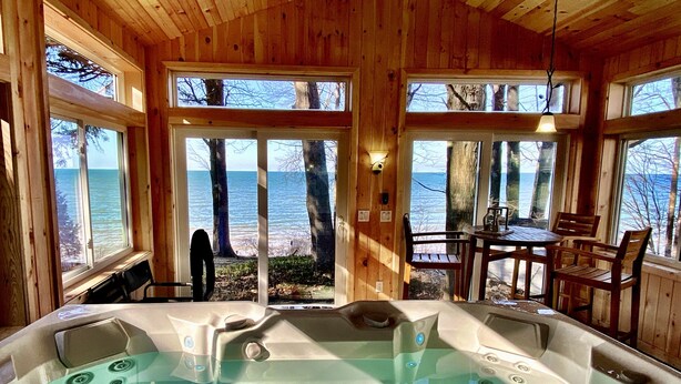 A wooden cabin with a hot tub, wooden furniture, and a view of the lake. - Lakefront Oak Cottage • Hot Tub & Fire Pit (Sodus)