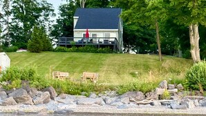 A lakeside house with a deck, a lawn, and a rocky shoreline. - Lakefront Oak Cottage • Hot Tub & Fire Pit (Sodus)