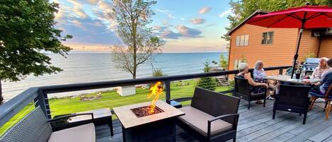 A deck with outdoor furniture and a fire pit, overlooking a body of water and a grassy area.